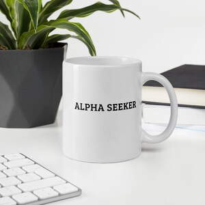 Può includere: Una tazza da caffè in ceramica bianca con una scritta nera che dice "ALPHA SEEKER".