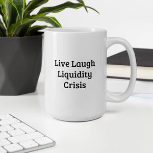 May include: A white ceramic mug with the words "Live Laugh Liquidity Crisis" printed in black. The mug has a large handle and is sitting on a white surface. A plant and a keyboard are in the background.