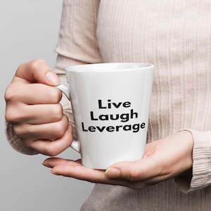 Könnte beinhalten: Eine weiße Keramik-Kaffeetasse mit schwarzem Text, der "Live Laugh Leverage" lautet.