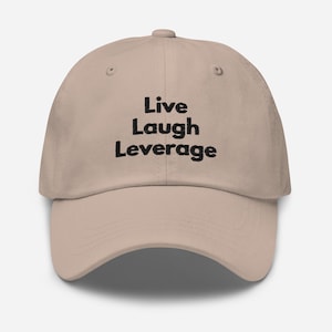 Op de afbeelding: Een beige baseballpet met zwarte geborduurde tekst die "Live Laugh Leverage" luidt.