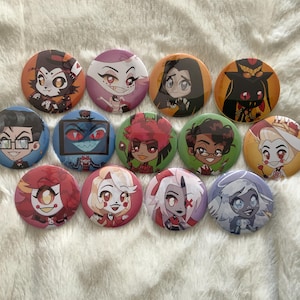 May include: A collection of eleven round pinback buttons featuring colorful cartoon character designs. The buttons are in various colors, including red, orange, blue, green, and purple. Each button has a unique character illustration.