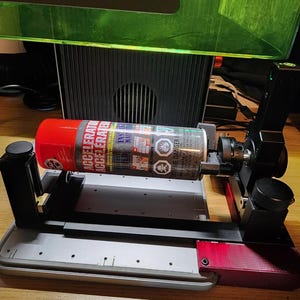 May include: A red and silver spray can labeled "ACCELERATED" is held in a black and gray machine. The machine is on a wooden surface, with a green cover in the background. A card with instructions is in the foreground.