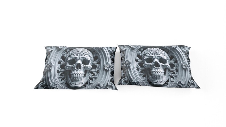 May include: Two decorative pillowcases with a detailed skull design. The skull is set within an ornate, circular frame with floral accents. The color palette is grayscale, with varying shades of gray.