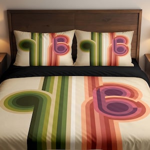 May include: Bedding set with a retro design featuring the letter "B" and curved stripes in green, orange, and purple. The set includes a duvet cover and two pillowcases on a cream background. The bed is made up with a black sheet.