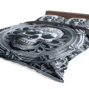 May include: A bed with a dark wooden headboard and a gray and white skull-themed bedding set. The duvet cover and pillowcases feature a detailed skull design within an ornate, floral-like frame. The overall aesthetic is gothic.