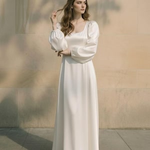 May include: A floor-length, off-white satin dress featuring a square neckline and long, voluminous puff sleeves with fitted cuffs. The garment has a smooth, flowing skirt that reaches the floor, creating a clean and simple silhouette.