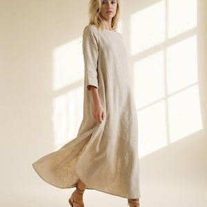 May include: A floor-length beige dress with three-quarter length sleeves, a round neckline, and a side slit, paired with brown strappy sandals. The garment features a loose, flowing silhouette and a textured fabric finish throughout the design.