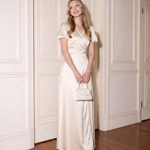 May include: A floor-length cream-coloured satin wrap dress with short puff sleeves and a V-neckline, paired with a small matching cream-coloured structured handbag and pointed-toe heels. The dress features a smooth, lustrous finish and a flowing skirt.