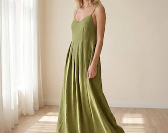 Puede incluir: Vestido largo verde oliva con cuello en V y tirantes finos. El vestido tiene un corpiño plisado y una falda fluida. La persona está de pie frente a una ventana con cortinas blancas, sobre un suelo de madera clara.