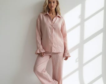 Puede incluir: Un conjunto de camisa de manga larga con botones y pantalón largo a juego en color rosa claro. La camisa presenta un cuello clásico y puños con volantes. El pantalón tiene un corte recto con dobladillos con volantes en la parte inferior.