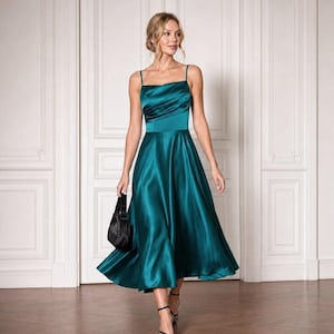 May include: A midi-length emerald green satin dress featuring a square neckline, thin spaghetti straps, a fitted bodice with subtle horizontal pleating, and a flared skirt. The dress has a smooth, reflective finish and a clean, structured silhouette.