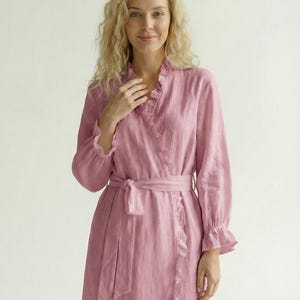 May include: A dusty rose wrap robe featuring long sleeves with ruffled cuffs, a V-neckline with ruffled trim along the front opening, and a matching fabric tie belt at the waist. The garment has a textured finish and falls to a midi length.