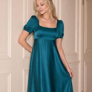 May include: A floor-length teal satin dress featuring a square neckline, short puff sleeves, and an empire waistline. The fabric has a smooth, lustrous finish that catches the light, and the skirt flows downward in a simple, unadorned silhouette.