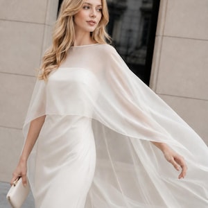 May include: A floor-length ivory gown featuring a strapless bodice and a sheer, flowing organza cape overlay that extends from the shoulders. The dress is paired with a small, rectangular, solid-colored clutch held in one hand.