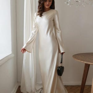 May include: A floor-length, long-sleeved cream satin dress with a round neckline and flared sleeves. The dress features a smooth, lustrous finish and a fluid, floor-sweeping hemline. A small, black, heart-shaped handbag is held at the side.