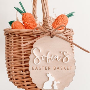 K&ouml;nnte beinhalten: Ein Weidenkorb mit einem Holzschild, auf dem "Sofia's Easter Basket" steht. Das Schild hat zwei wei&szlig;e Hasenschnitte. Der Korb ist mit orangefarbenen Karotten-Dekorationen und braunem Stroh gef&uuml;llt.