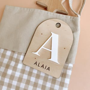 May include: A beige and white gingham stocking with a wooden tag that says "ALAIA" and has a white letter "A" on it. The tag is attached to the stocking with a brown string.