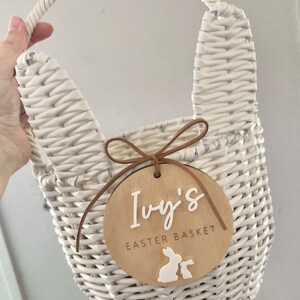 K&ouml;nnte beinhalten: Ein wei&szlig;er Weidenkorb mit Hasenohren und einem Holzschild mit der Aufschrift "Ivy's Easter Basket" und zwei wei&szlig;en Hasensilhouetten.