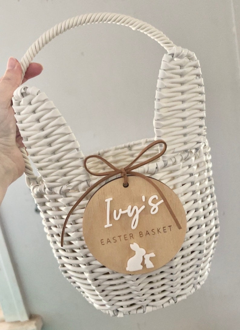 K&ouml;nnte beinhalten: Ein wei&szlig;er Weidenkorb mit Hasenohren und einem Holzschild mit der Aufschrift "Ivy's Easter Basket" und zwei wei&szlig;en Hasensilhouetten.
