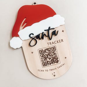 May include: A wooden Santa Tracker ornament shaped like a Santa hat. The red hat has a white trim and the word "Santa" in black script. Below is a QR code and the text "SCAN TO TRACK SANTA!"