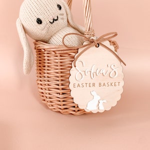 K&ouml;nnte beinhalten: Ein Weidenkorb mit einem wei&szlig;en gestrickten Hasen darin. Der Korb hat ein Holzschild mit der Aufschrift "Sofia's Easter Basket" und zwei wei&szlig;en Hasensilhouetten.