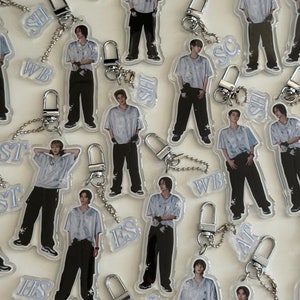 Puede incluir: Una colección de 12 llaveros de acrílico transparente con diferentes imágenes de hombres jóvenes. Cada llavero tiene una cadena plateada y un cierre plateado. Los llaveros están decorados con texto de diferentes colores, incluyendo azul, blanco y negro.