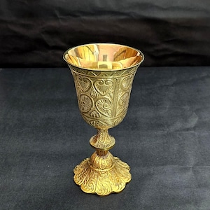Könnte beinhalten: Ein goldfarbener Metallkelch mit einem verzierten Design. Der Kelch hat eine breite, ausgestellte Basis und einen hohen, schlanken Stiel.