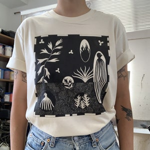 Peut inclure: Un t-shirt blanc avec un imprimé graphique noir et blanc de plantes et d'oiseaux stylisés. Le graphique est encadré d'une bordure à carreaux noir et blanc.