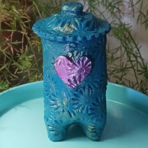 May include: A teal blue ceramic jar with a lid and a purple heart design. The jar has a textured surface with a floral pattern. The jar has three legs.