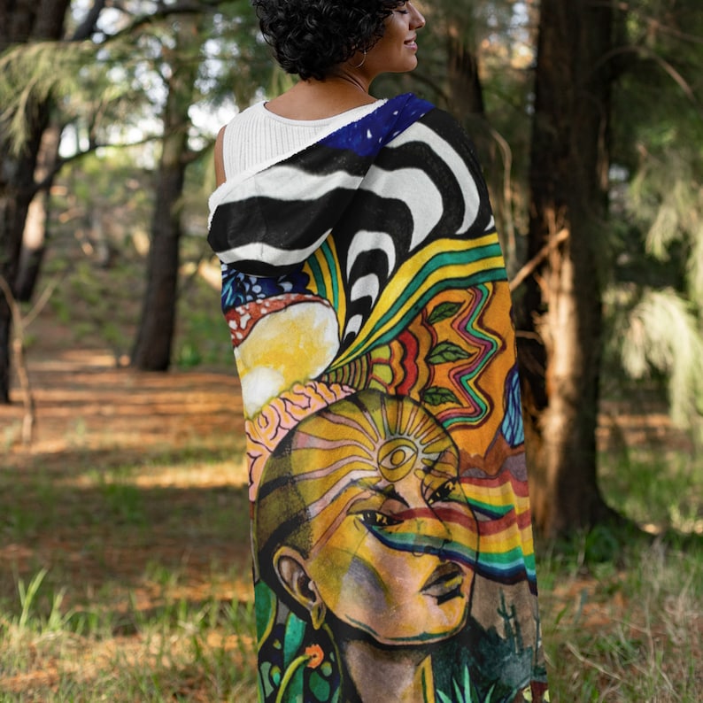 May include: A colorful, artistic blanket featuring a vibrant illustration. The design includes a portrait with a third eye, rainbow streaks, and psychedelic patterns in shades of yellow, blue, green, and orange. The blanket has a black and white striped pattern on the hood.