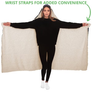 May include: A hooded blanket with a cream-colored, fluffy interior and a dark blue exterior trim. The blanket has wrist straps for added convenience. The person in the image is wearing a black sweater and black leggings.