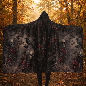 May include: A hooded cloak featuring a dark, intricate pattern of human skulls, black crows perched on branches, and deep red roses. The fabric displays a moody, gothic-inspired print with grey and black foliage set against a dark background.