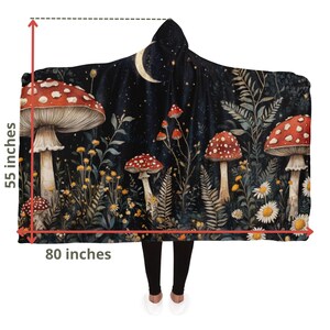 Puede incluir: Una manta con capucha que presenta una escena de bosque caprichosa con setas rojas y blancas, follaje verde y una luna creciente contra un cielo nocturno estrellado azul oscuro. La manta mide 140 cm por 203 cm.