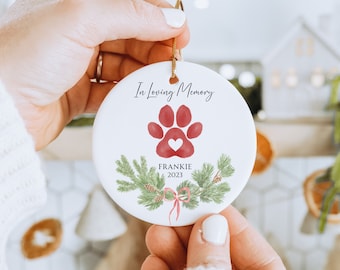 May include: White ceramic Christmas ornament with a red paw print and a heart in the center. The ornament has green pine branches and a red bow. The text "In Loving Memory Frankie 2023" is printed on the ornament.