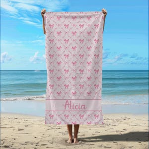 May include: A pink beach towel with a white background and a pattern of pink bows. The towel has the name "Alicia" printed on it in pink.