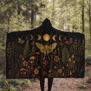 Op de afbeelding: Zwarte capuchonponcho met een op de natuur geïnspireerd ontwerp. De poncho heeft een grote mot, halve manen, vlinders, paddenstoelen en bloemelementen in goud, groen en bruin. De achtergrond is een bosscène.