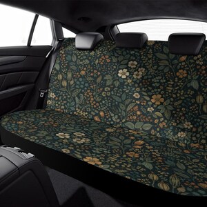 May include: Car seat cover with a dark background and a floral pattern in shades of green, yellow, and cream. The design features various flowers and leaves, creating a botanical theme. The cover is designed to fit the back seat of a vehicle.