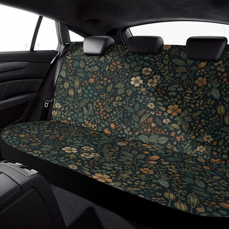 May include: Car seat cover with a dark background and a floral pattern in shades of green, yellow, and cream. The design features various flowers and leaves, creating a botanical theme. The cover is designed to fit the back seat of a vehicle.