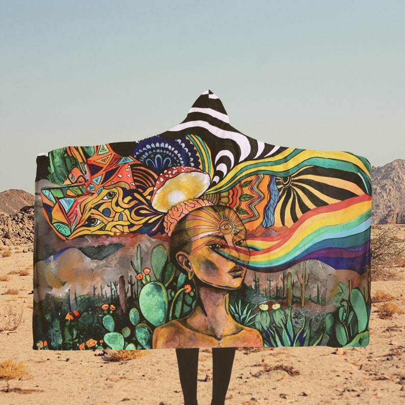 May include: A hooded blanket featuring a vibrant, surreal illustration. The design includes a woman's face with a rainbow emanating from her eye, a desert landscape, and colorful psychedelic patterns. The hood has a black and white zebra stripe pattern.