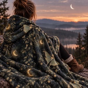 May include: A hooded blanket featuring a dark teal background with a repeating pattern of pale green moths, golden crescent moons, small stars, and delicate leafy branches. The fabric has a soft, plush texture and covers the shoulders and back.
