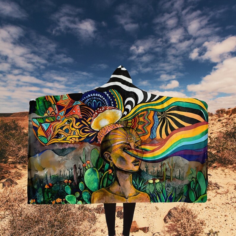 May include: A hooded blanket featuring a vibrant, surreal illustration. The design includes a woman's face with a rainbow emanating from her nose, surrounded by colorful patterns, cacti, and a desert landscape. The hood has a black and white zebra stripe pattern.