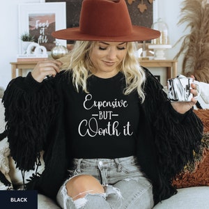 May include: A black t-shirt with the text "Expensive -BUT- Worth it" in white lettering. The model is wearing the shirt with a brown hat and jeans.