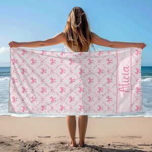 May include: A pink beach towel with a white background and a pattern of pink bows. The towel has a white border with the name "Alicia" written in pink.