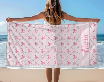 May include: A pink beach towel with a white background and a pattern of pink bows. The towel has a white border with the name "Alicia" written in pink.