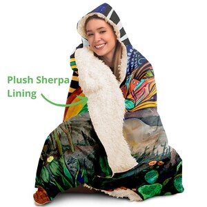 May include: A hooded blanket with a colorful, artistic design. The blanket features a plush sherpa lining for added warmth and comfort. The design includes a variety of colors and patterns, creating a unique and eye-catching look.