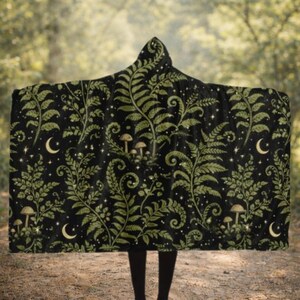 May include: A hooded blanket featuring a black background with a repeating pattern of green fern fronds, small brown mushrooms, and yellow crescent moons interspersed with tiny white stars across the entire fabric surface.