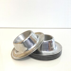 May include: A set of silver-toned metal egg cups with a polished finish. One egg cup is tilted and resting against a stack of three identical egg cups. Each piece features a wide circular base and a central cylindrical holder for an egg.