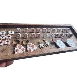 May include: A wooden tray displays various handmade earrings. The earrings feature a variety of designs, including crosses, geometric shapes, and floral patterns. The color palette includes white, brown, and gold, with some earrings having a textured appearance.