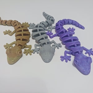 May include: Three articulated lizard figurines in shades of tan, gray, and purple. Each lizard has a segmented body, four legs, and a head with detailed features. The lizards are arranged on a white surface, showcasing their unique colors and designs.