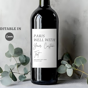 Op de afbeelding: Een zwarte wijnfles met een wit etiket met de tekst "Paris Well With Your Custom Text". Het etiket bevat ook de tekst "Add more text here if you'd like". Eucalyptustakken staan op de voorgrond.